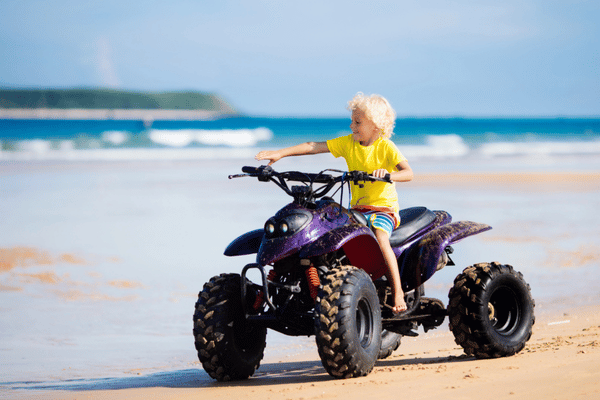 Young child rides an ATV across wet sand by the ocean on a sunny day.
