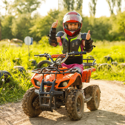 Child wearing full protective gear gives thumbs up while riding ATV on a dirt path