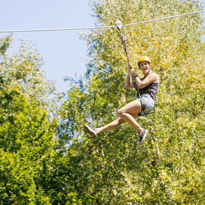 A smiling woman rides a zipline through trees, an exciting reminder to ask is ziplining safe