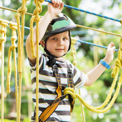 A child wearing a helmet and harness navigates a ropes course with yellow climbing ropes safely