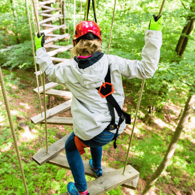 Participant crosses forest suspension bridge, confronting fear of heights, wearing helmet, gloves, and safety harness