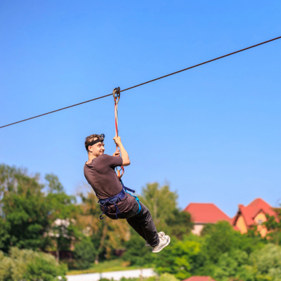 Man enjoys an outdoor ziplining experience, gliding above trees and houses against a clear blue sky