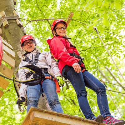 Two harnessed friends on treetop platform smile confidently after completing challenging high-rope adventure course today