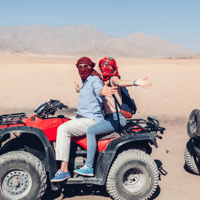 Couple on red ATV pose arms wide in desert landscape with mountains and bright sky