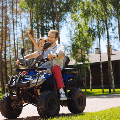 Smiling atv family rides blue quad near cabins, child waving excitedly through sunny birch trees