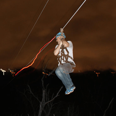 Rider speeds along night zipline, harness secure, lights streaking across dark sky during thrilling adventure