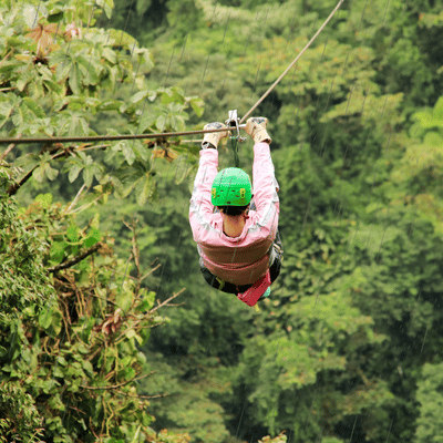 Rider faces forward on rainforest zipline during light rain, wearing helmet, gloves, and pink jacket